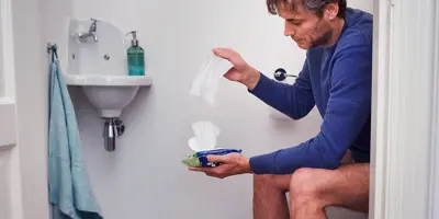 Ein junger Mann sitzt auf einer Toilette und hält eine Taschentuchbox in seinen Händen.