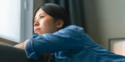 Eine junge Frau in einem Jeanshemd schaut gedankenverloren aus einem Fenster in die Ferne.