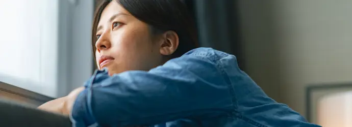 Eine junge Frau in einem Jeanshemd schaut gedankenverloren aus einem Fenster in die Ferne.