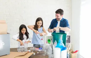 Una famiglia felice sorride mentre schiaccia bottiglie di plastica e appiattisce un cartone per il riciclo.