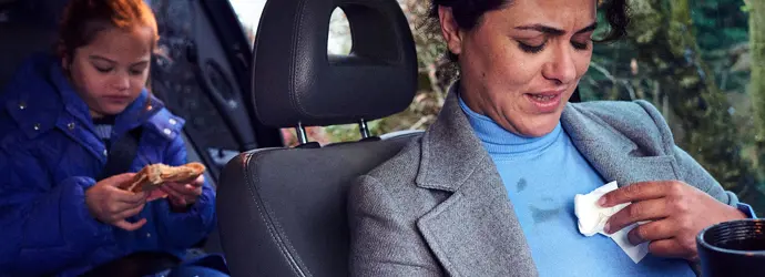 Een vrouw zit op de voorstoel van een auto haar blouse schoon te maken; een meisje eet een boterham op de achterbank.