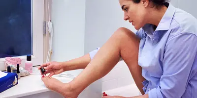 Een vrouw lakt haar teennagels in een badkamer; een doos tissues ligt voor haar.