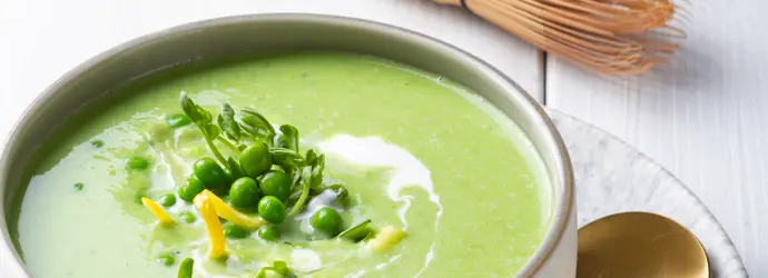 Grüne Matcha-Erbsensuppe garniert mit Erbsen und Zitronenschale in einer Schüssel. Daneben ein goldener Löffel und ein Matcha Besen.