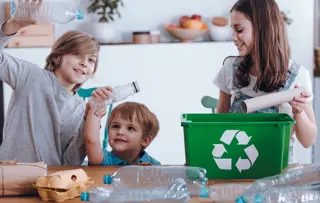 Tre fratelli sorridono mentre selezionano bottiglie di plastica, cartoni delle uova e tubi di cartone da riciclare.