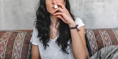 Eine junge Frau mit Birkenpollenallergie sitzt auf einem Sofa und benutzt ein Nasenspray