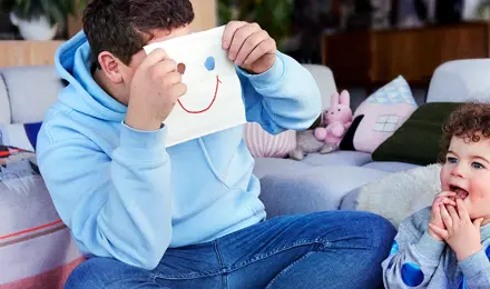 Ein Mann sitzt mit einem Kind auf dem Boden und hält ein Taschentuch mit einem Smiley vor seinem Geischt in die Höhe.