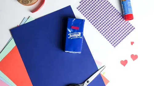 Bastelsachen auf weißem Tisch: buntes Papier, Tempo-Taschentücher, Schere, Klebestift, Glitzersteine und Herzchen.