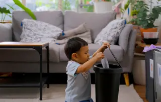 Bambino con bavaglino a pois che mette i rifiuti di plastica trasparente in un bidone nero a casa.