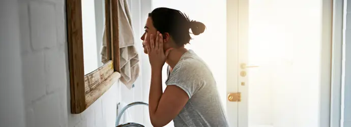 Eine Frau steht vor einem Spiegel in einem Badezimmer und hält ihre rechte Hand an die rechte Seite ihres Gesichts und betrachtet sich im Spiegel.