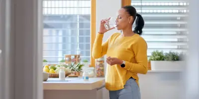 Eine Frau in einem gelben Pullover steht in einer sonnigen Küche und trinkt ein Glas Wasser.