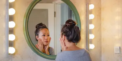 Una ragazza castana con i capelli raccolti si guarda allo specchio e distende il viso con le mani.