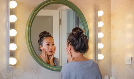 Una ragazza castana con i capelli raccolti si guarda allo specchio e distende il viso con le mani.