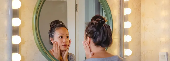 Una ragazza castana con i capelli raccolti si guarda allo specchio e distende il viso con le mani.