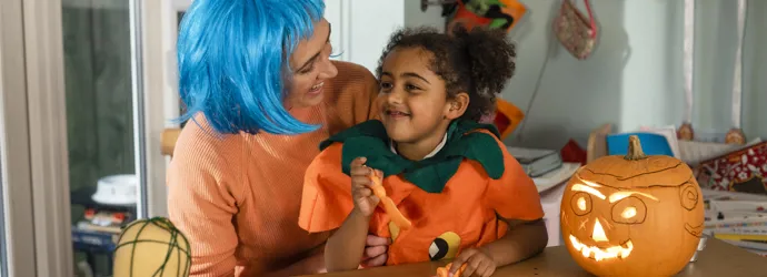 Ein verkleidetes Kind auf dem Schoß einer erwachsenen Person, umgeben von selbst gebastelter Halloween Deko.