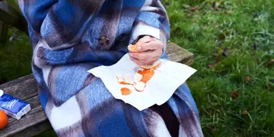 Een oudere vrouw in een geruite jas zit op een bankje in het park en eet een sinaasappel om haar immuunsysteem te versterken.