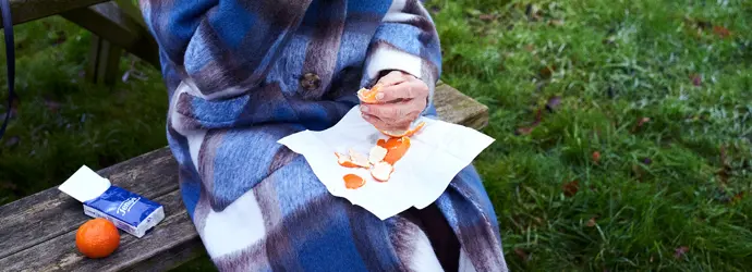 Een oudere vrouw in een geruite jas zit op een bankje in het park en eet een sinaasappel om haar immuunsysteem te versterken.