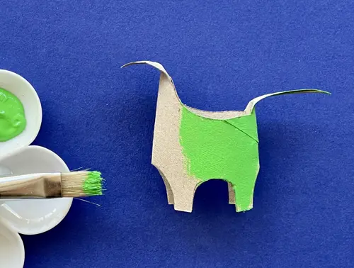 Dinosauro fatto con rotolo di carta igienica in parte dipinto di verde. Accanto un pennello su una tavolozza con del verde.