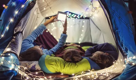 Drei Kinder liegen bei einer Übernachtungsparty nebeneinander in einem geschmückten und beleuchteten Zelt.