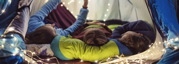 Drei Kinder liegen bei einer Übernachtungsparty nebeneinander in einem geschmückten und beleuchteten Zelt.