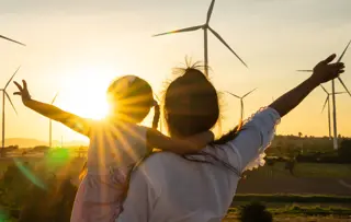 Una madre e un bambino di fronte al tramonto, a braccia aperte, con turbine eoliche sullo sfondo.