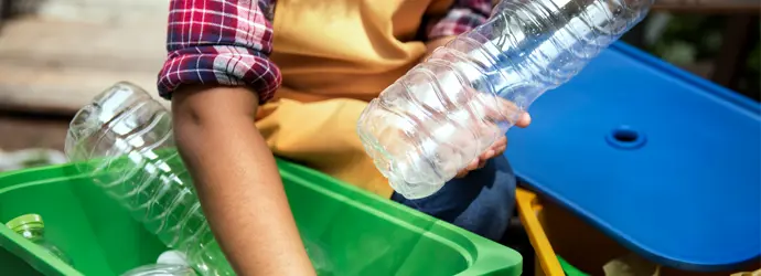 Un bambino con una camicia a quadri che smista bottiglie di plastica in un bidone verde per il riciclaggio all'aperto.