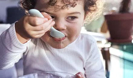 Een peuter met donker krullend haar eet yoghurt met een lepel uit een glazen schaaltje.
