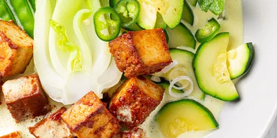 Knusprig gebratene Tofu Würfel neben grünem Gemüse, Chilischoten und Kokoscurry auf einem weißen Porzellanteller.