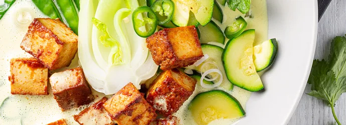 Knusprig gebratene Tofu Würfel neben grünem Gemüse, Chilischoten und Kokoscurry auf einem weißen Porzellanteller.