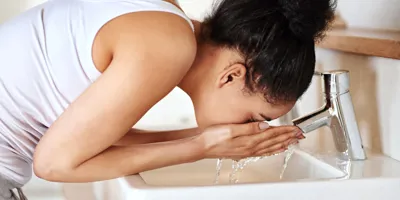 Una donna castana con i capelli in uno chignon e canotta bianca è china sul lavandino del bagno e si sciacqua il viso.