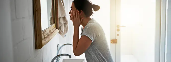 Donna con t-shirt grigia si tocca il viso e si osserva nello specchio del bagno.