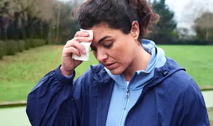 Een vrouw veegt het zweet van haar voorhoofd terwijl ze met griep in een park loopt.
