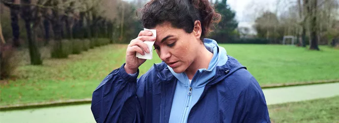 Een vrouw veegt het zweet van haar voorhoofd terwijl ze met griep in een park loopt.