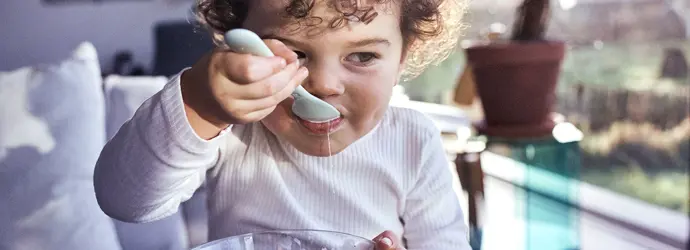 Un bambino con i capelli ricci e scuri mangia lo yogurt con un cucchiaio da una piccola ciotola di vetro.