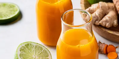 Zwei Gläschen mit orangenem Ingwer-Kurkuma-Shot neben Limettenscheiben und einer Schale mit frischen Kurkumawurzeln.