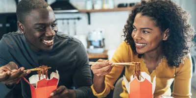 Ein junger Mann und eine junge Frau sitzen an einem Tisch, essen Nudeln aus einer Pappverpackung und sehen sich an.