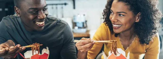 Ein junger Mann und eine junge Frau sitzen an einem Tisch, essen Nudeln aus einer Pappverpackung und sehen sich an.