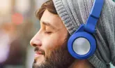 Uomo con barba e felpa con cappuccio grigio che ascolta musica con cuffie blu e occhi chiusi.