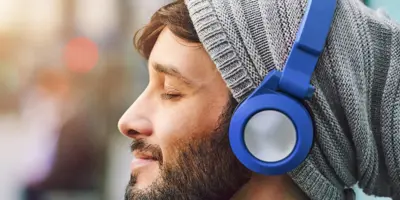 Uomo con barba e felpa con cappuccio grigio che ascolta musica con cuffie blu e occhi chiusi.