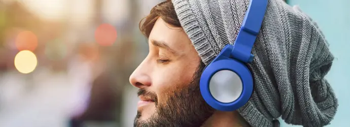 Uomo con barba e felpa con cappuccio grigio che ascolta musica con cuffie blu e occhi chiusi.