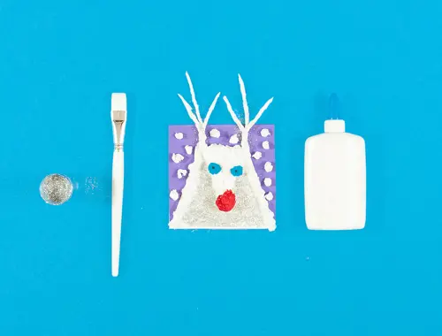 Un ritaglio al centro con volto e orecchie di una renna con accanto un pennello e contenitore bianco e glitter argentati.