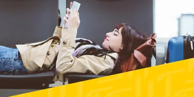 Una donna bruna è sdraiata su due sedie in un aeroporto, leggendo un libro mentre aspetta il suo volo. Accanto a lei c'è una valigia blu.
