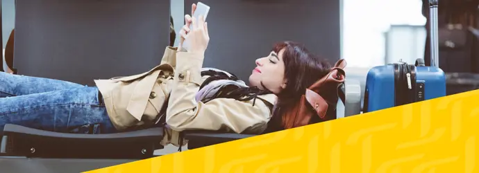 Una donna bruna è sdraiata su due sedie in un aeroporto, leggendo un libro mentre aspetta il suo volo. Accanto a lei c'è una valigia blu.