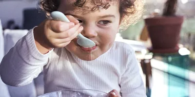 Ein Kleinkind mit lockigen dunklen Haaren isst Joghurt mit einem Löffel aus einer kleinen Glasschüssel.
