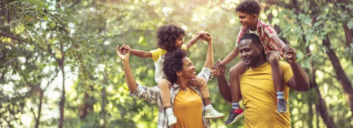 Una famiglia sorridente si gode una passeggiata nella foresta. I genitori portano i figli sulle spalle.
