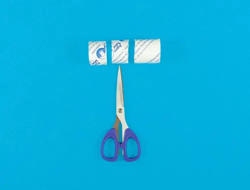 Un piccolo rotolo di carta igienica tagliato in tre sezioni giace su uno sfondo blu con in basso un paio di forbici a punta.