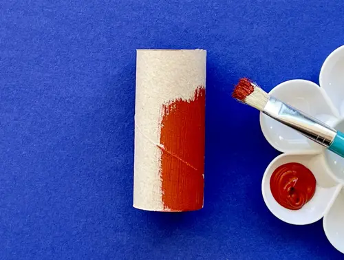 Su sfondo blu un pennello su una tavolozza con del colore rosso. Accanto un rotolo di carta igiencia dipinto di rosso.