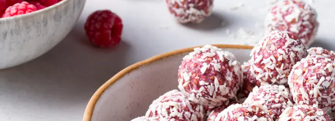 Mehrere pinke Himbeer-Energy-Balls mit Kokosraspeln in einer hellen Schüssel. Im Hintergrund steht eine Schüssel mit Himbeeren.