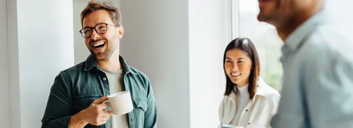 Zwei Männer und eine Frau stehen in einem Raum und lächeln, während alle eine Tasse in der Hand halten.