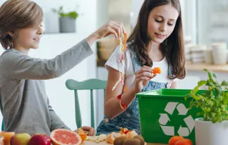 Due bambini smistano bucce di frutta e scarti di cibo in un bidone verde per il riciclaggio.