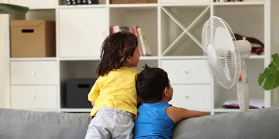 Due bambini si rinfrescano con un ventilatore affacciati allo schienale di un divano grigio.